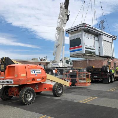 A crane lifts a small building, construction vehicles and workers are around
