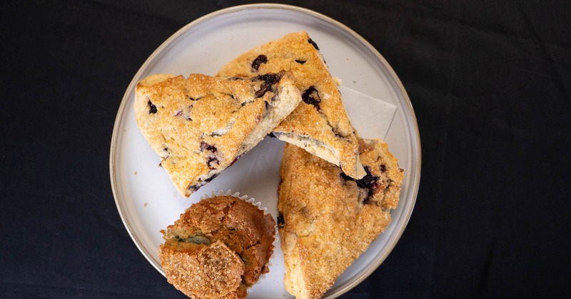 Blueberry scones and a muffin on a white plate, set on a dark table