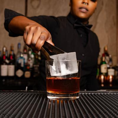 Bartender using tongs to place a large, clear ice cube into a cocktail glass.