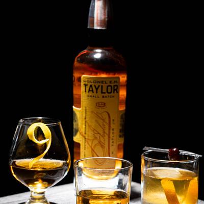 A bottle of whiskey stands behind three glasses of whiskey with orange peels, dark background.