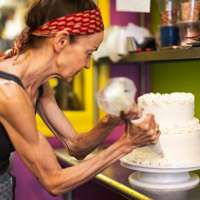 An employee  decorates a two-tier cake with white frosting using a piping bag.