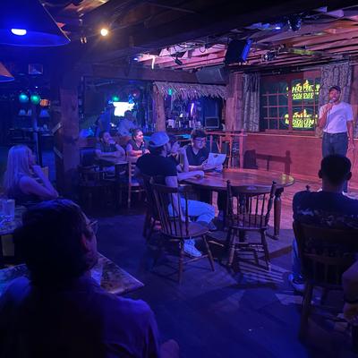 An audience watches a comedian on stage, seated at wooden tables