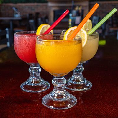 Three colorful frozen cocktails in schooner glasses with straws and lemon slices.