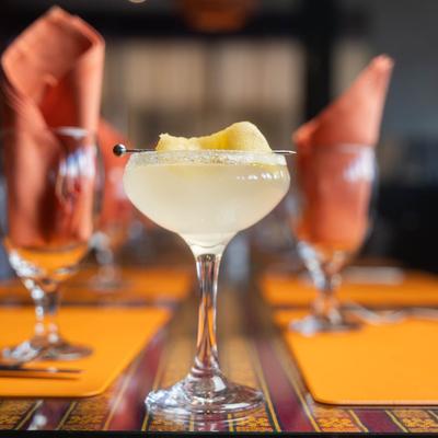 A yellow drink, garnished with lemon peel, sits on a table with folded orange napkins.