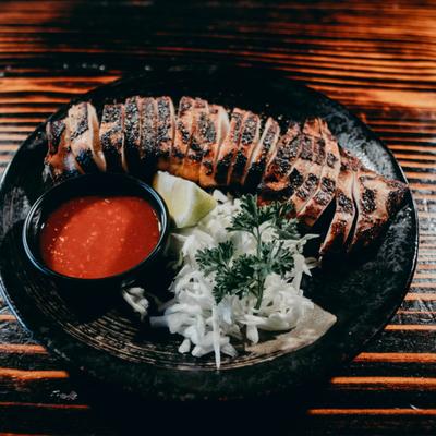 Grilled squid served on a black plate with a side of red sauce, lemon wedge, and cabbage garnish.