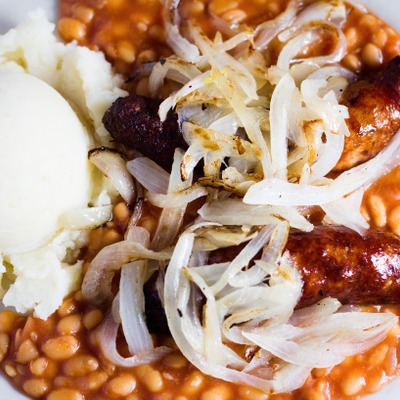 Banger sausages topped with grilled onions on top of baked beans and next to mashed potatoes