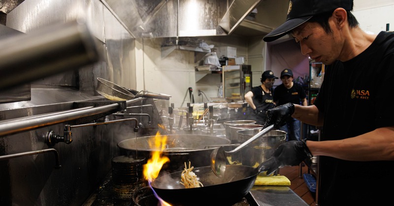 Person cooking noodles in a wok on a professional stovetop