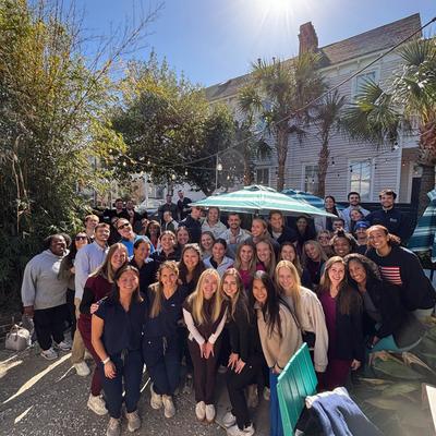 A group of smiling people poses  under a bright sun, surrounded by greenery and palm trees.