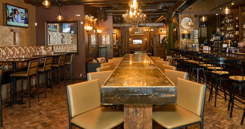 Rustic bar interior with a long wooden table and beige chairs. Exposed ducts and industrial lights.