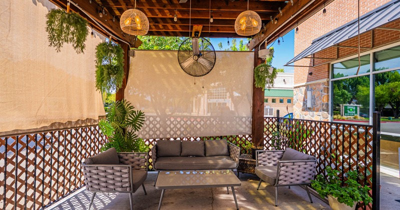 Covered patio with wicker seating, plants, hanging lights, fabric shades, and a large ceiling fan