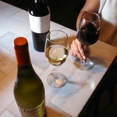 Two glasses of wine on a marble table with bottles nearby.