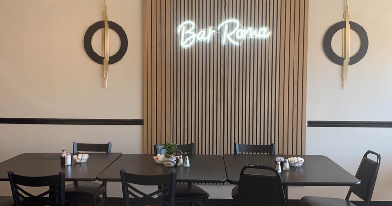 Dining area with a neon "Bar Roma" sign, black tables and modern wall decor.