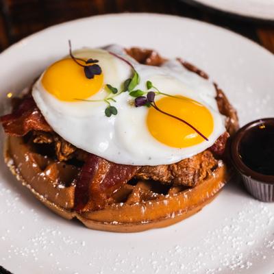 Belgian waffle topped with fried chicken, bacon, two fried eggs, and microgreens.