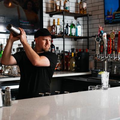 A bartender shaking a cocktail with beer taps and liquor bottles visible in the background.