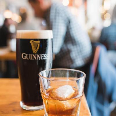 A pint of Guinness and a whiskey glass with ice sit on a wooden table in a lively pub.
