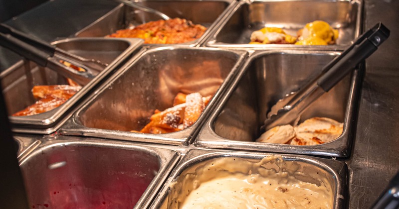 Buffet food service line featuring stainless steel steam table pans containing various dishes