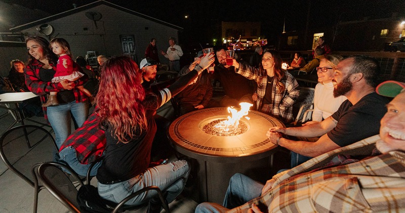 Nighttime in the patio, customers sitting around a fire pit table and toasting with drinks