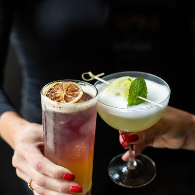 Woman in black holds two cocktails, a lime-green gin cocktail and a two-tone honey-chacha and pineapple cocktail