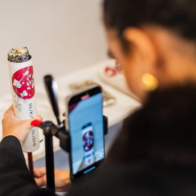 Person is using a smartphone on a stand to photograph the sushi roll in the tube.