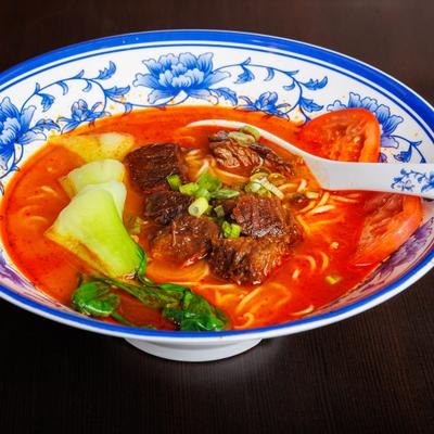 Spicy beef noodle soup with bok choy, tomatoes, and scallions n a blue floral bowl.
