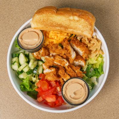 A salad bowl - lettuce, tomatoes, cucumbers, cheddar cheese, chicken pieces, toast, dressing cups.