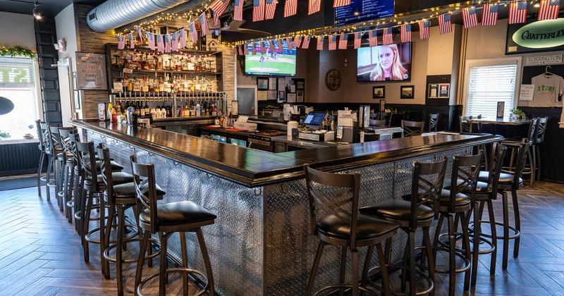 Large metal-fronted bar with several high-top stools and multiple TVs