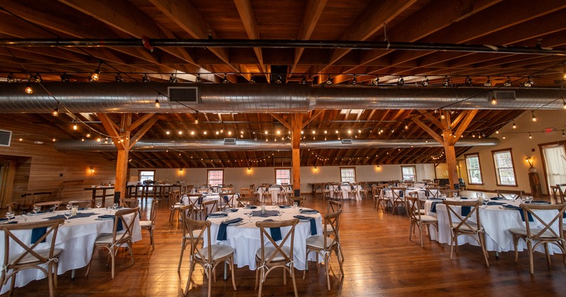 A rustic event hall - wooden beams, string lights, set with white-clothed tables and wooden chairs
