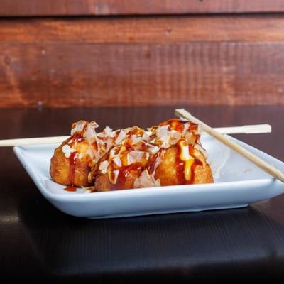 Takoyaki drizzled with sauces and bonito flakes served on a white plate with chopsticks.
