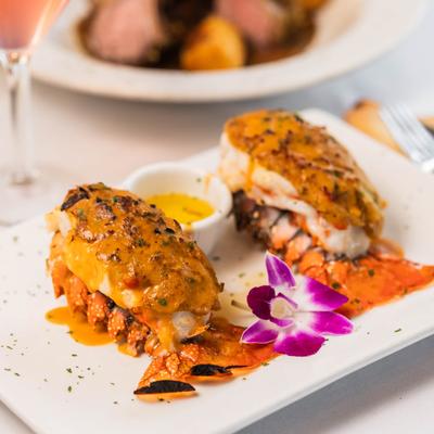 Twin lobster tails on a white plate with drawn butter cup and purple flower garnish.