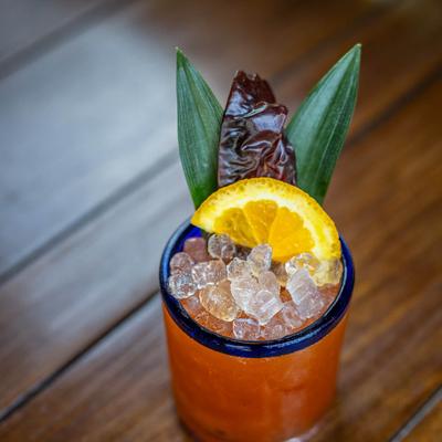 A craft red cocktail with crushed ice, an orange slice, and green leaves.
