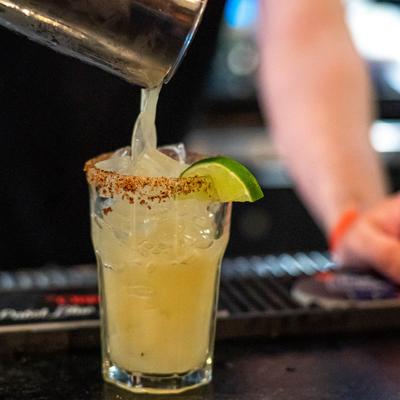 Bartender pouring a light-yellow cocktail into a glass, garnished with spiced rim and a lime wedge.