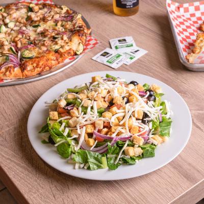 Garden salad with shredded cheese and croutons on a table with a pizza nearby.