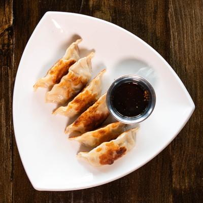 Potstickers and a metal cup of soy sauce arranged on a white plate, atop wooden table.