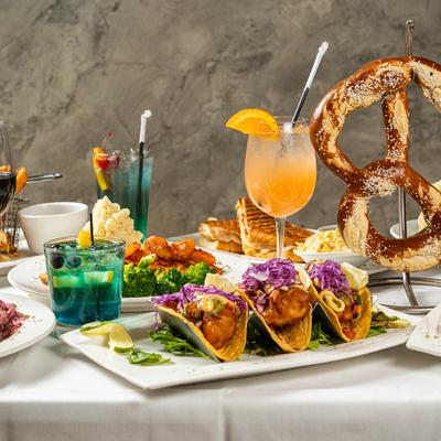 Various dishes and drinks on a table, including fish tacos, sandwiches, and a pretzel.