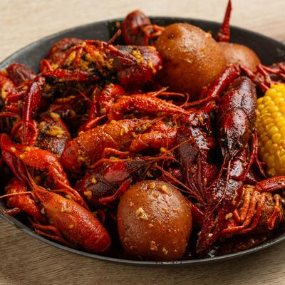 Black skillet with red crawfish, two  potatoes, and a corn cob on a wooden table.
