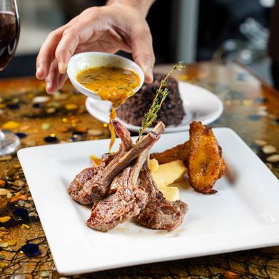 A hand pouring sauce on lamb chops with sides on a plate.