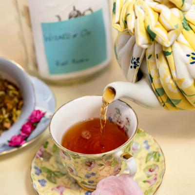 Porcelain teapot with a yellow floral cozy pouring amber tea into a matching teacup.