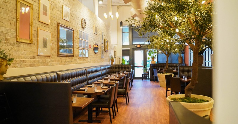 Interior - leather booths, wooden tables, potted trees and framed artwork on a brick wall.