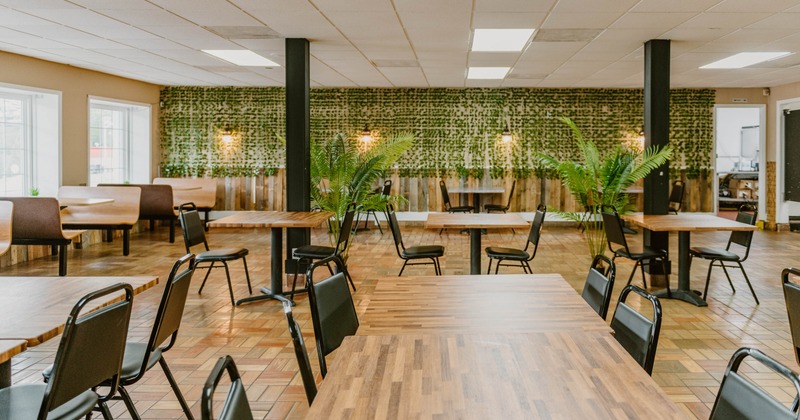 A spacious restaurant interior with wooden tables, black chairs, booth seating, and a green foliage wall