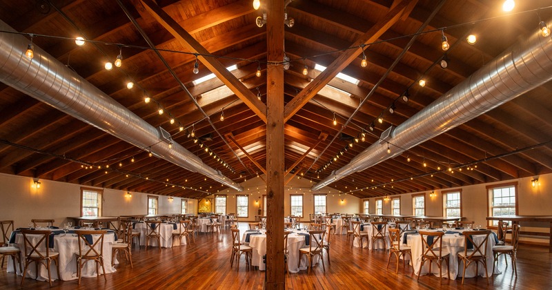 A rustic banquet hall - wooden beams, string lights, round tables set with white tablecloths.