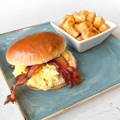 A breakfast sandwich on a brioche bun with scrambled eggs and bacon, served with potatoes.