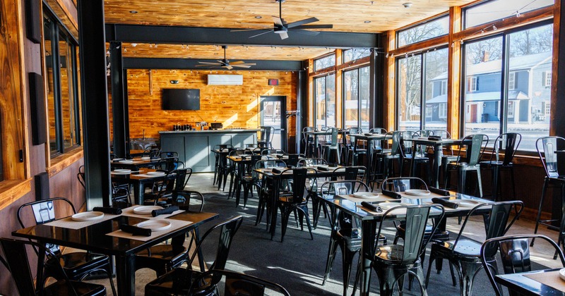 Bright restaurant interior with wooden ceiling, black tables and metal chairs, and large windows
