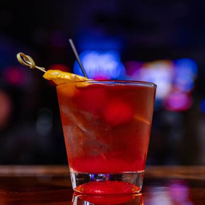 Red cocktail with lemon slice and cherry garnish, rests on a bar counter.