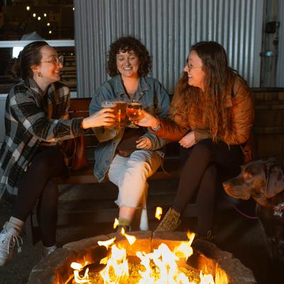 People outside sitting around a fire pit toasting with their drinks.