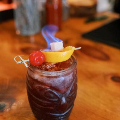Red cocktail garnished with a cherry, lemon slice, and a flaming sugar cube.