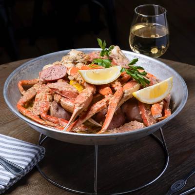 A steaming platter of crab legs, sausage, potatoes, and corn, a glass of white wine sits beside on a wooden table.
