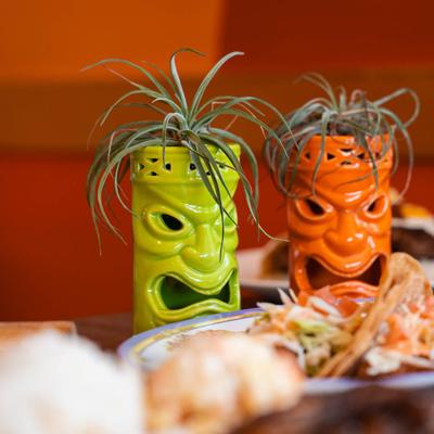 Two  tiki planters, one green and one orange, with plants on top, adorn a dining table.