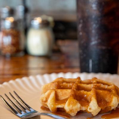 A waffle drizzled with a caramel-colored sauce, served on a white paper plate with a fork.