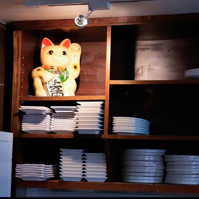Maneki-neko figurine sits on a wooden shelf above stacks of white plates.