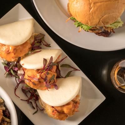 Top down view, a plate of Bao Sliders next to a burger and a glass of whiskey.